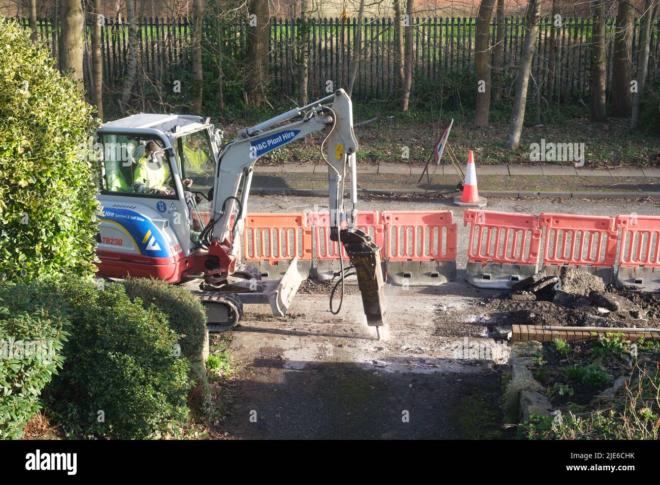 Workman using a mini excavator to dig up a pavement (sidewalk) by a road in a residential area Stock Photo
