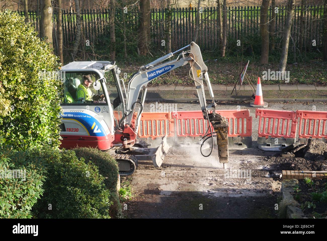 Workman using a mini excavator to dig up a pavement (sidewalk) by a road in a residential area Stock Photo