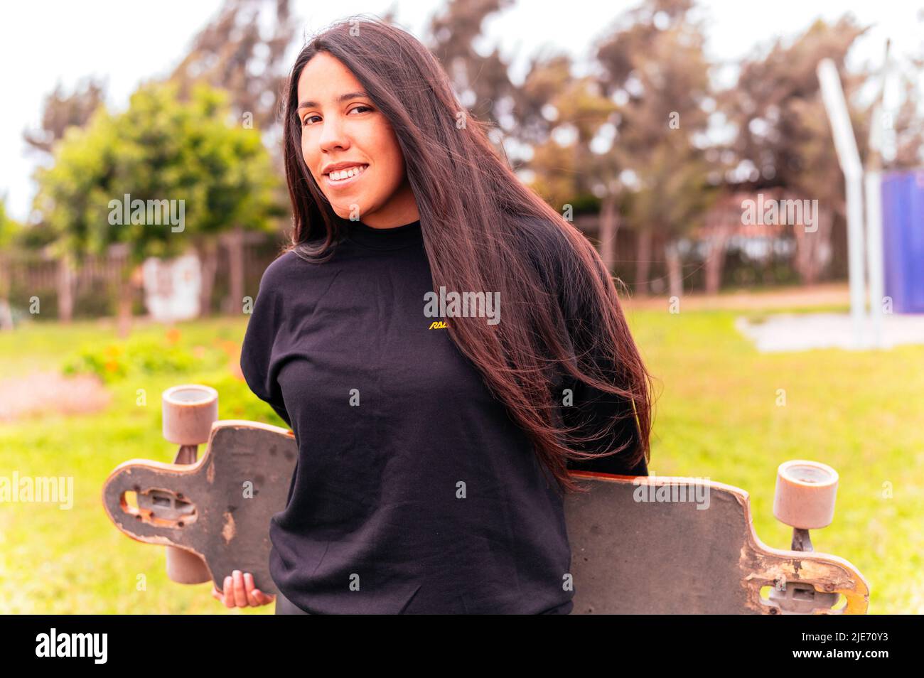 Portrait of smiling young female skateboarder holding her skateboard. Woman with skating board at skate park looking at camera outdoors Stock Photo