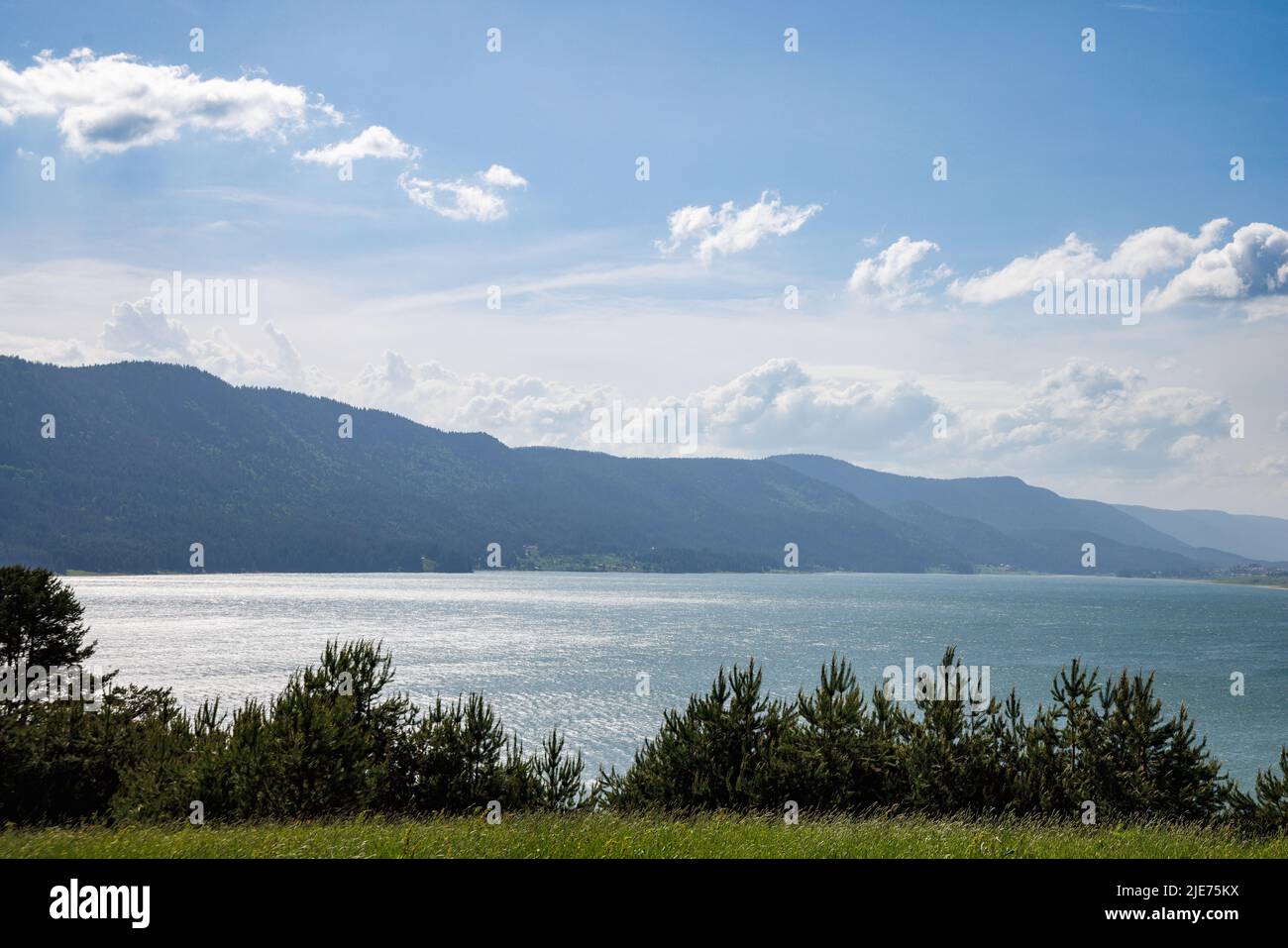 A gleaming wide clear lake reflects bright warm sunlight, against a clear blue cloudy sky and rolling green wooded mountain ranges and small farming m Stock Photo