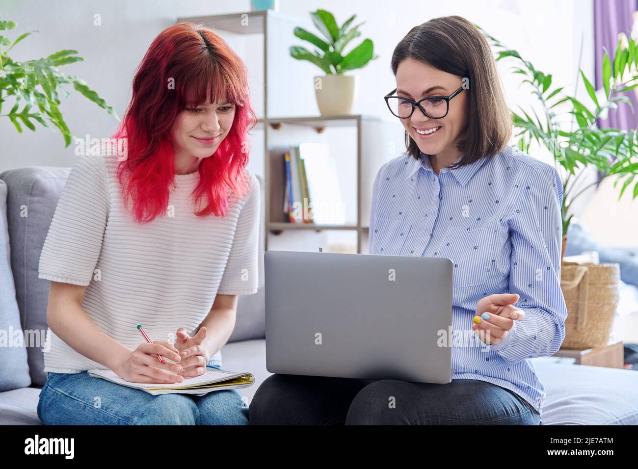 Female teacher teaches teenage girl, sitting together on couch in office, using laptop Stock
