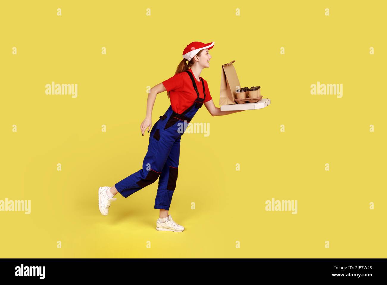 Side view portrait of delivery woman being hurry to deliver coffee with pizza, fast delivery service, wearing overalls and red cap. Indoor studio shot isolated on yellow background. Stock Photo
