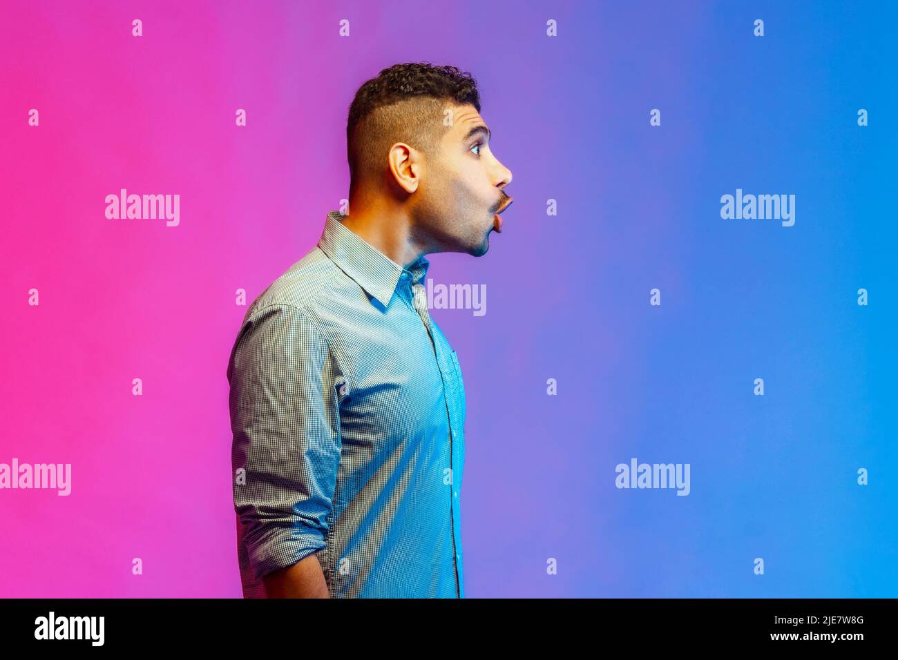 Portrait of astonished man in shirt looking right with big eyes and open mouth, shocked by news, empty copy space for text. Indoor studio shot isolated on colorful neon light background. Stock Photo