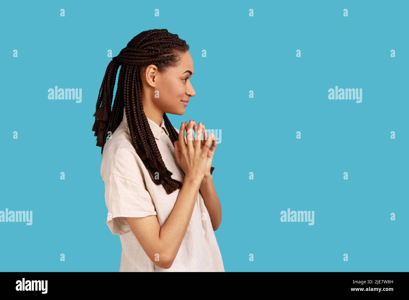 Side view of thoughtful woman with black dreadlocks schemes something, keeps fingers together, considers over cunning plans, wearing white shirt. Indoor studio shot isolated on blue background. Stock Photo