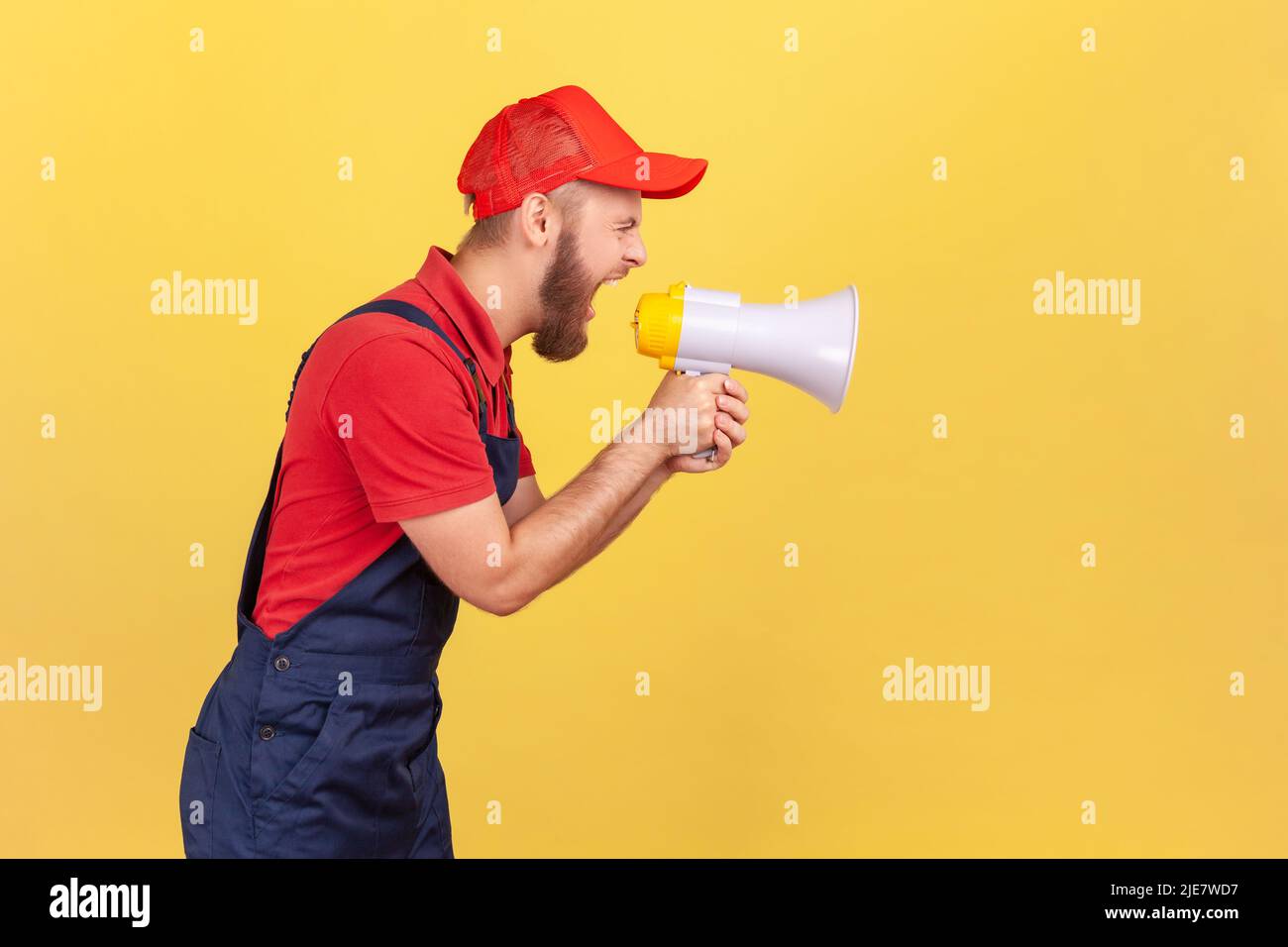Side view portrait of angry bearded worker in blue uniform and red cap, holding megaphone and screaming with aggressive expression, protesting. Indoor studio shot isolated on yellow background. Stock Photo