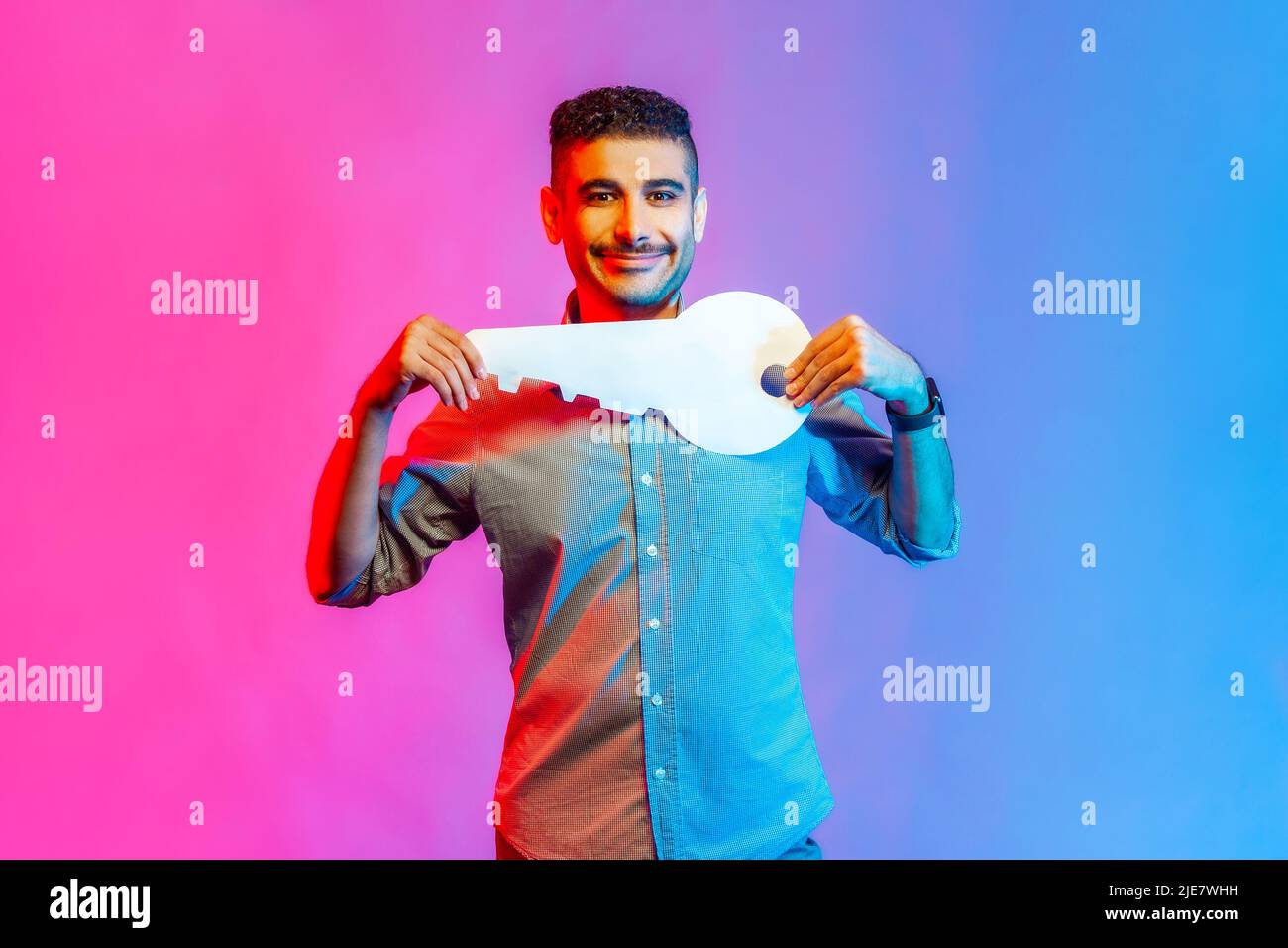Portrait of positive man in shirt holding huge paper key and smiling satisfied with new home, real estate purchase, rental service. Indoor studio shot isolated on colorful neon light background. Stock Photo