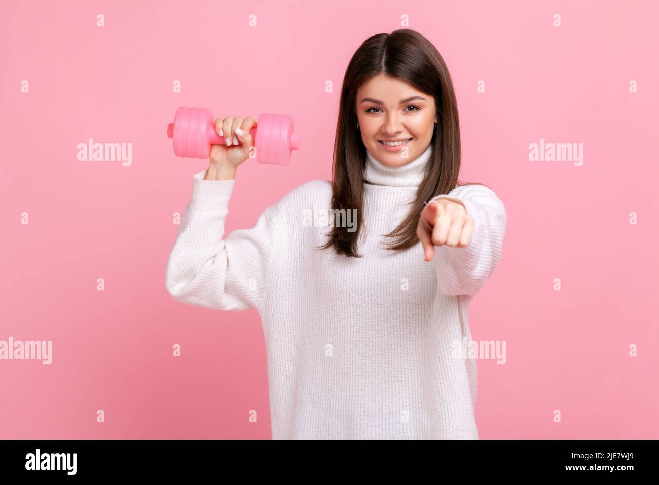 Smiling female holding rose dumbbells and pointing finger to camera, calls on to go in for sport, wearing white casual style sweater. Indoor studio shot isolated on pink background. Stock Photo