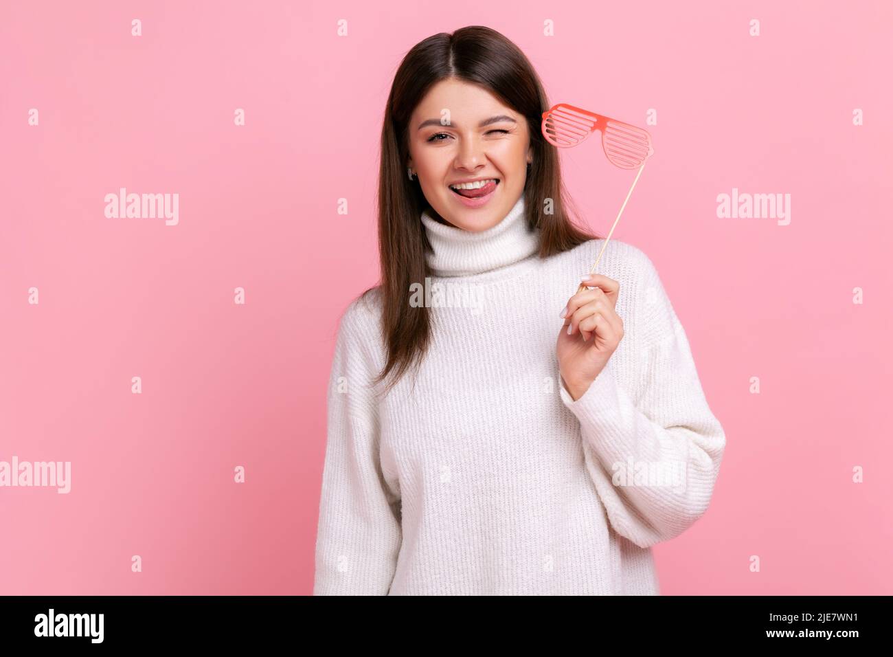 Happy playful girl looking at camera and winking, holding paper glasses, party props for celebration, wearing white casual style sweater. Indoor studio shot isolated on pink background. Stock Photo