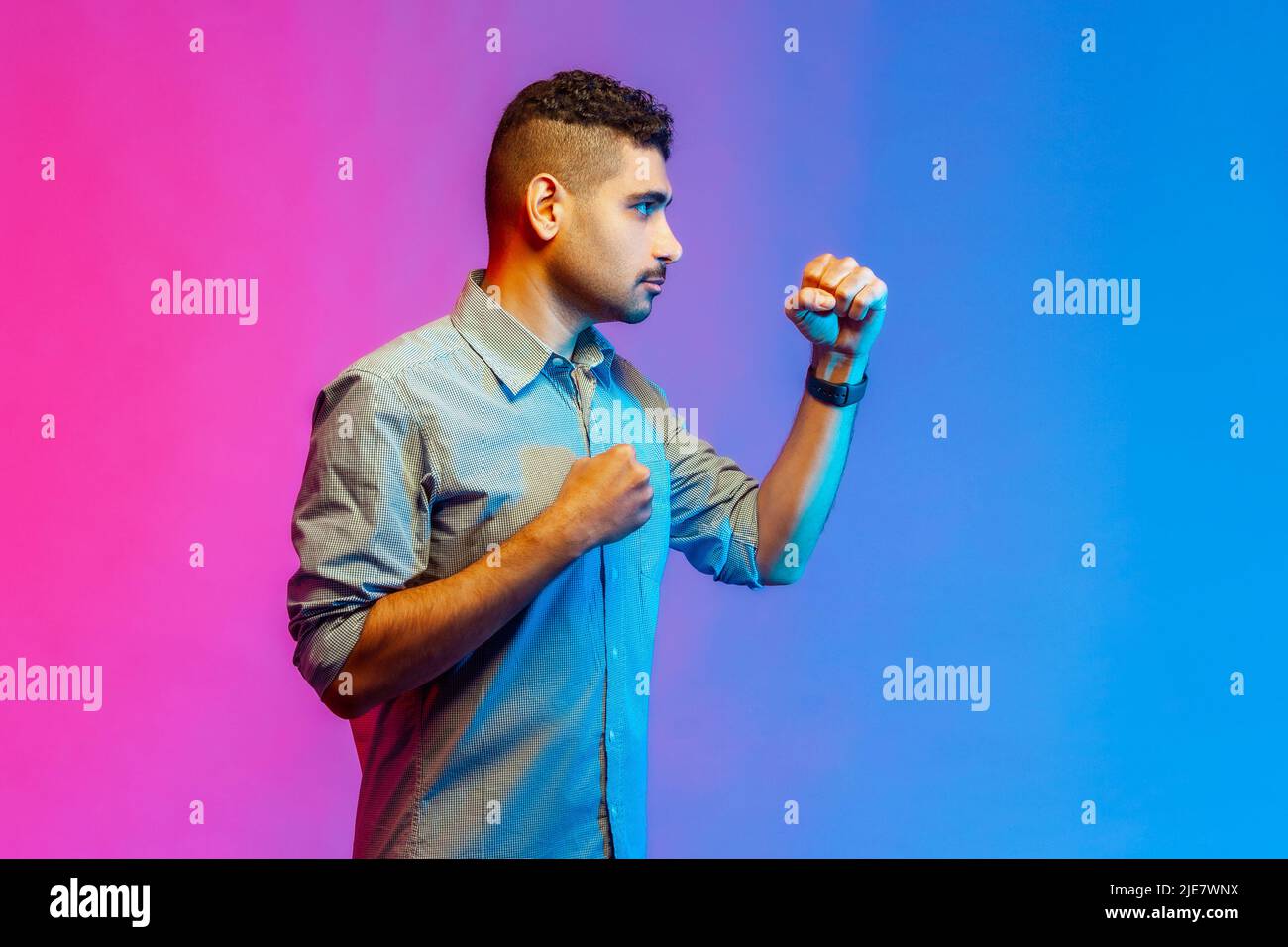 Side view portrait of young adult angry aggressive man in shirt punching, boxing with clenched fists, being ready fighting. Indoor studio shot isolated on colorful neon light background. Stock Photo