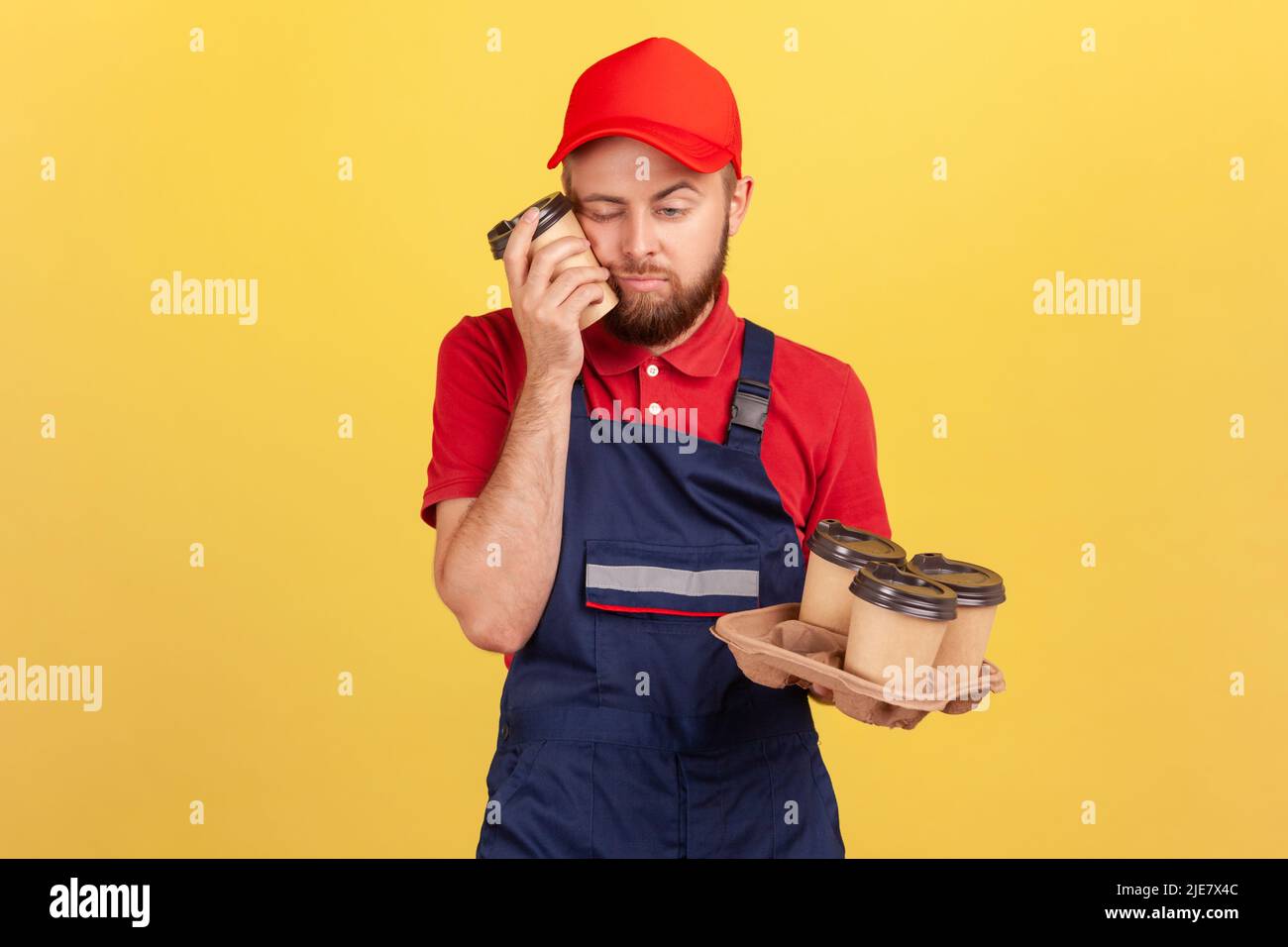 Tired exhausted worker feels sleepy after hard work standing with closed eyes, having nap, holding disposable cups of coffee, wearing overalls and cap. Indoor studio shot isolated on yellow background Stock Photo