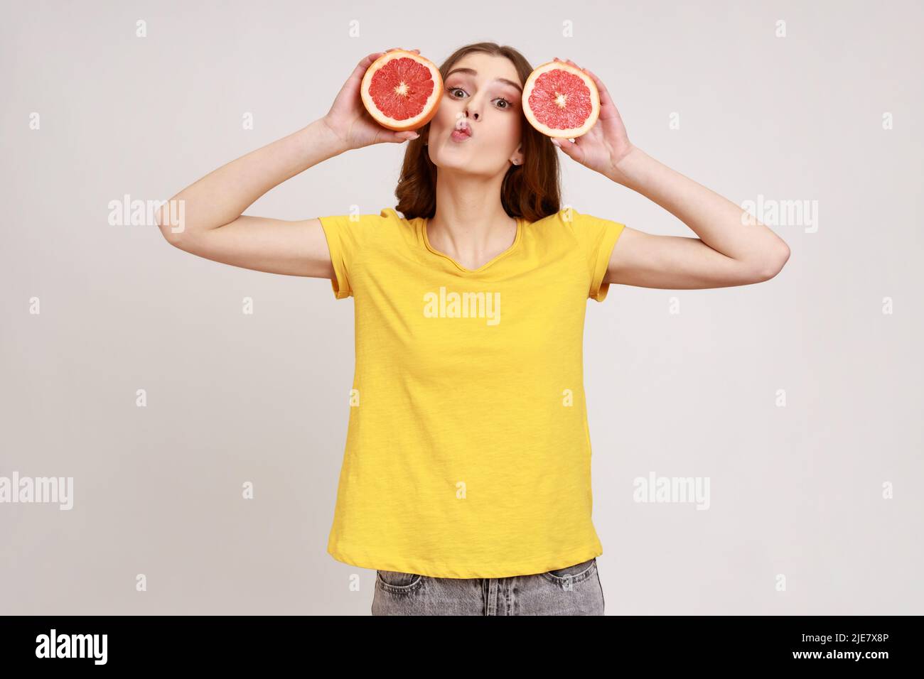 Portrait of positive teenager girl with dark hair holding two half slices of grapefruit with funny expression, making kissing gesture, looking at camera. Indoor studio shot isolated on gray background Stock Photo