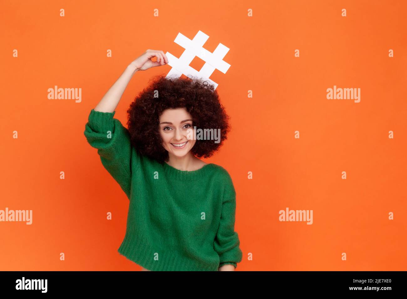 Portrait of funny positive woman with Afro hairstyle wearing green casual style sweater standing holding hashtag above her head, smiling. Indoor studio shot isolated on orange background. Stock Photo