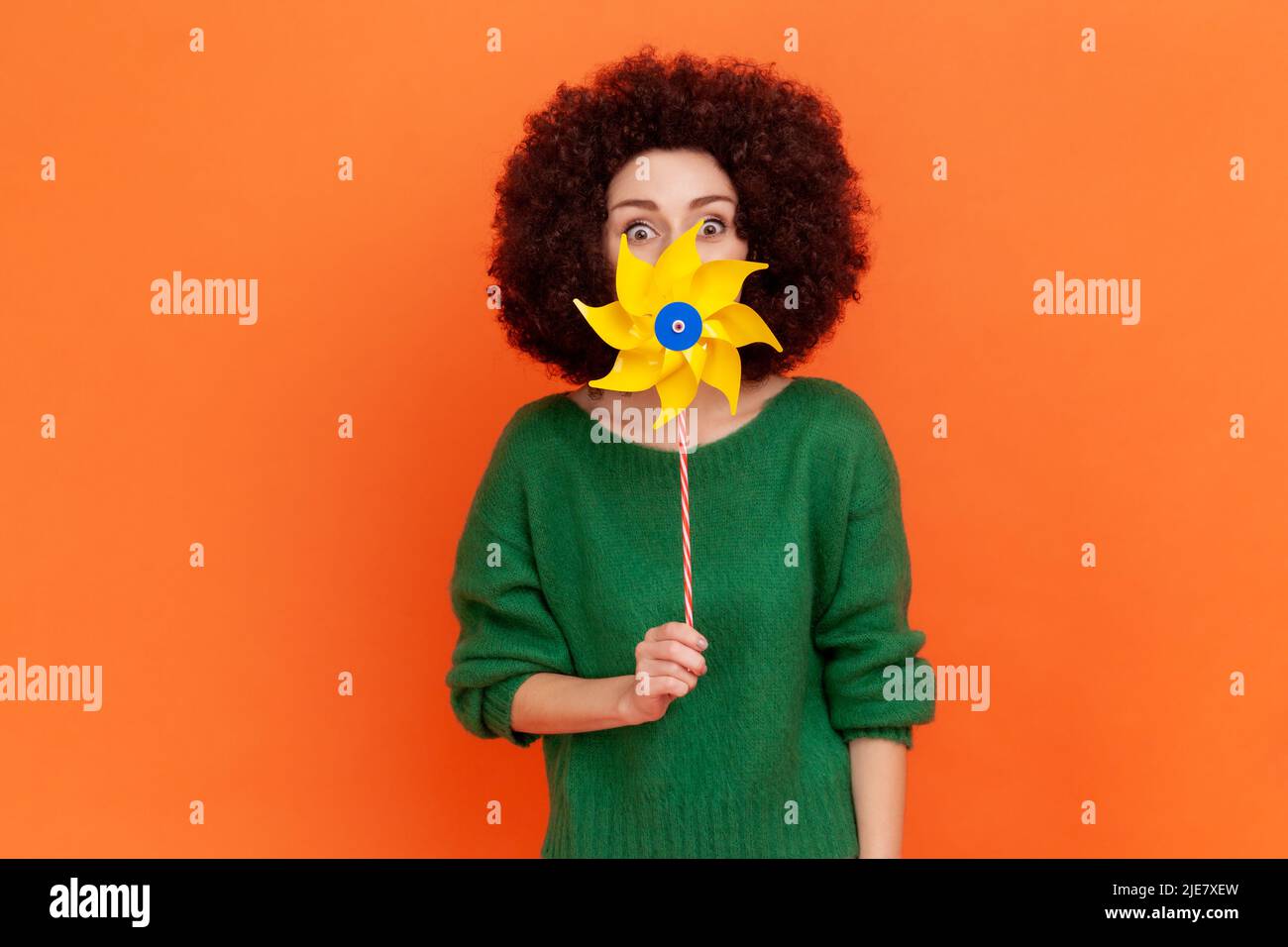 Attractive funny woman with Afro hairstyle wearing green casual style sweater hiding half of her face with yellow windmill, playing, having fun. Indoor studio shot isolated on orange background. Stock Photo
