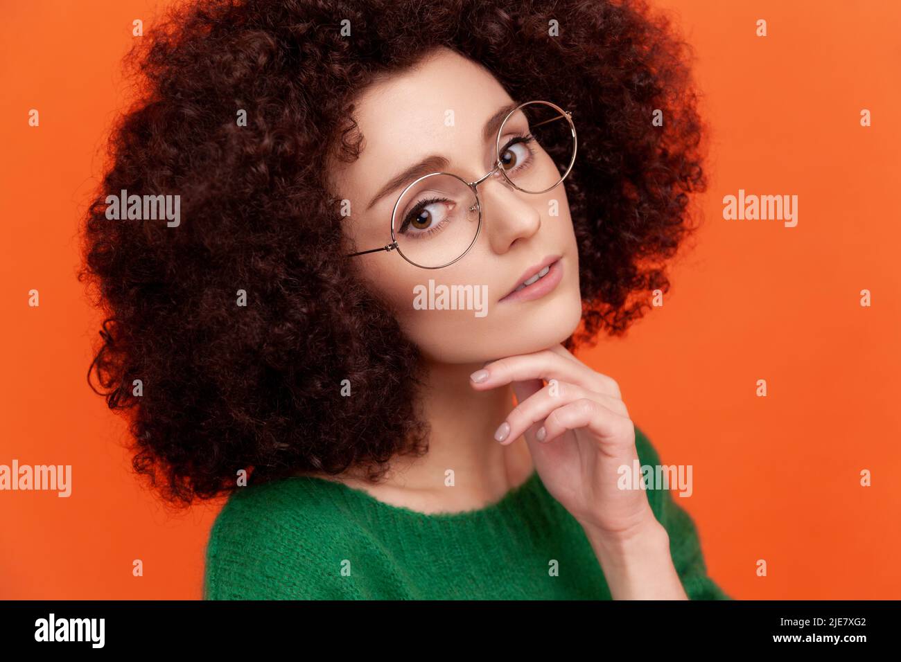 Closeup portrait of pleasant looking woman with Afro hairstyle wearing green casual style sweater and eyeglasses, keeping hand under chin. Indoor studio shot isolated on orange background. Stock Photo