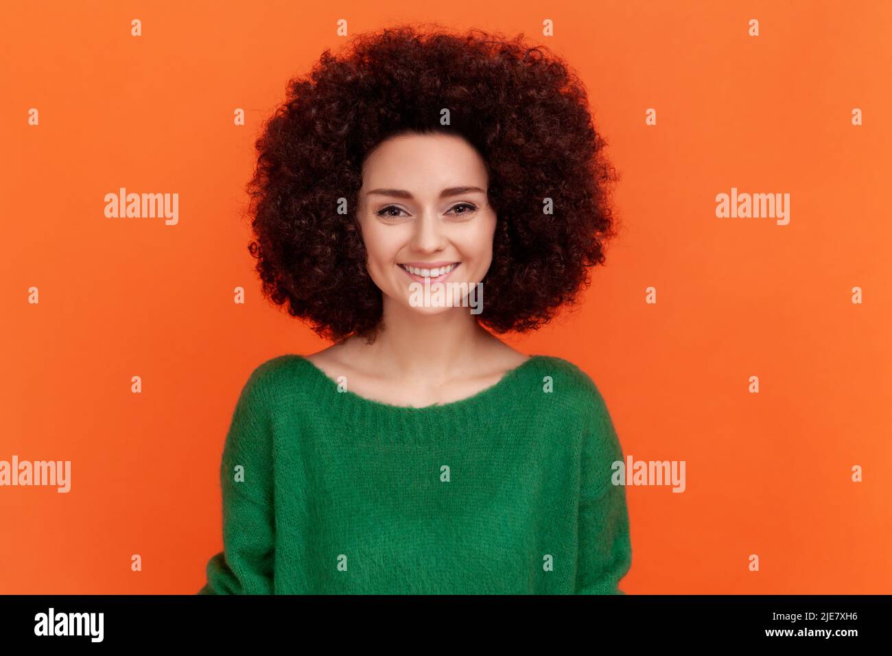 Portrait of beautiful positive woman with Afro hairstyle wearing green casual style sweater looking at camera, has pleasant emotions. Indoor studio shot isolated on orange background. Stock Photo