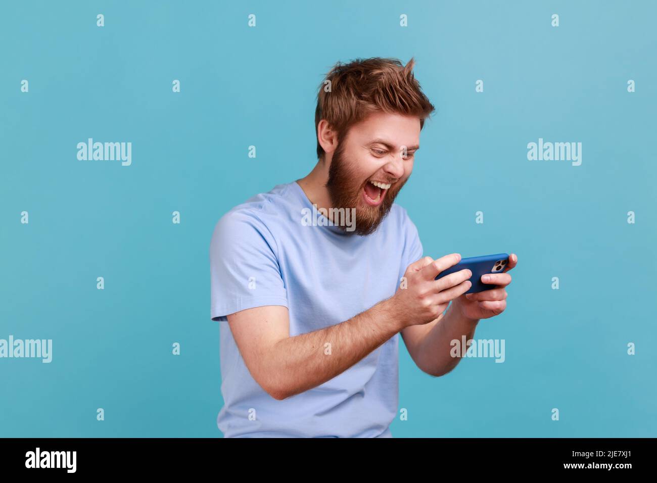 Portrait of handsome young adult bearded man standing, using smartphone and playing mobile game with excited positive face, happy to win. Indoor studio shot isolated on blue background. Stock Photo