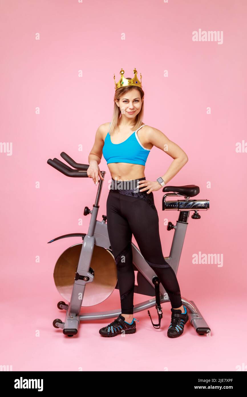 Full length of athletic woman in golden crown, queen of sports trainings, standing with hand on hips near bike simulator, wearing sports tights and top. Indoor studio shot isolated on pink background. Stock Photo