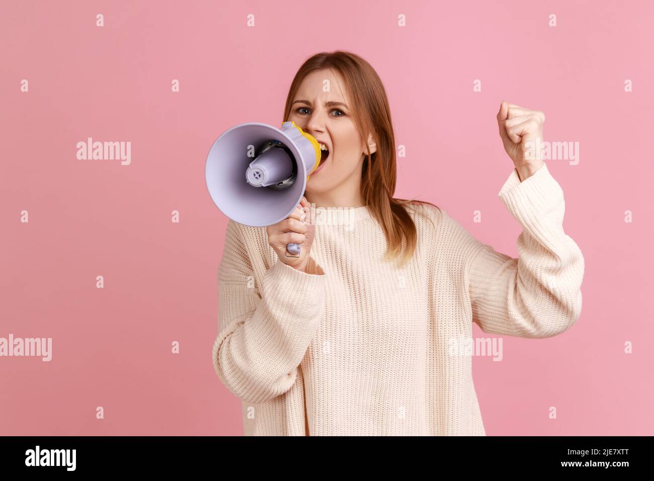 Portrait of serious concentrated blond woman protesting, raised arm and screaming in megaphone, looking at camera, wearing white sweater. Indoor studio shot isolated on pink background. Stock Photo
