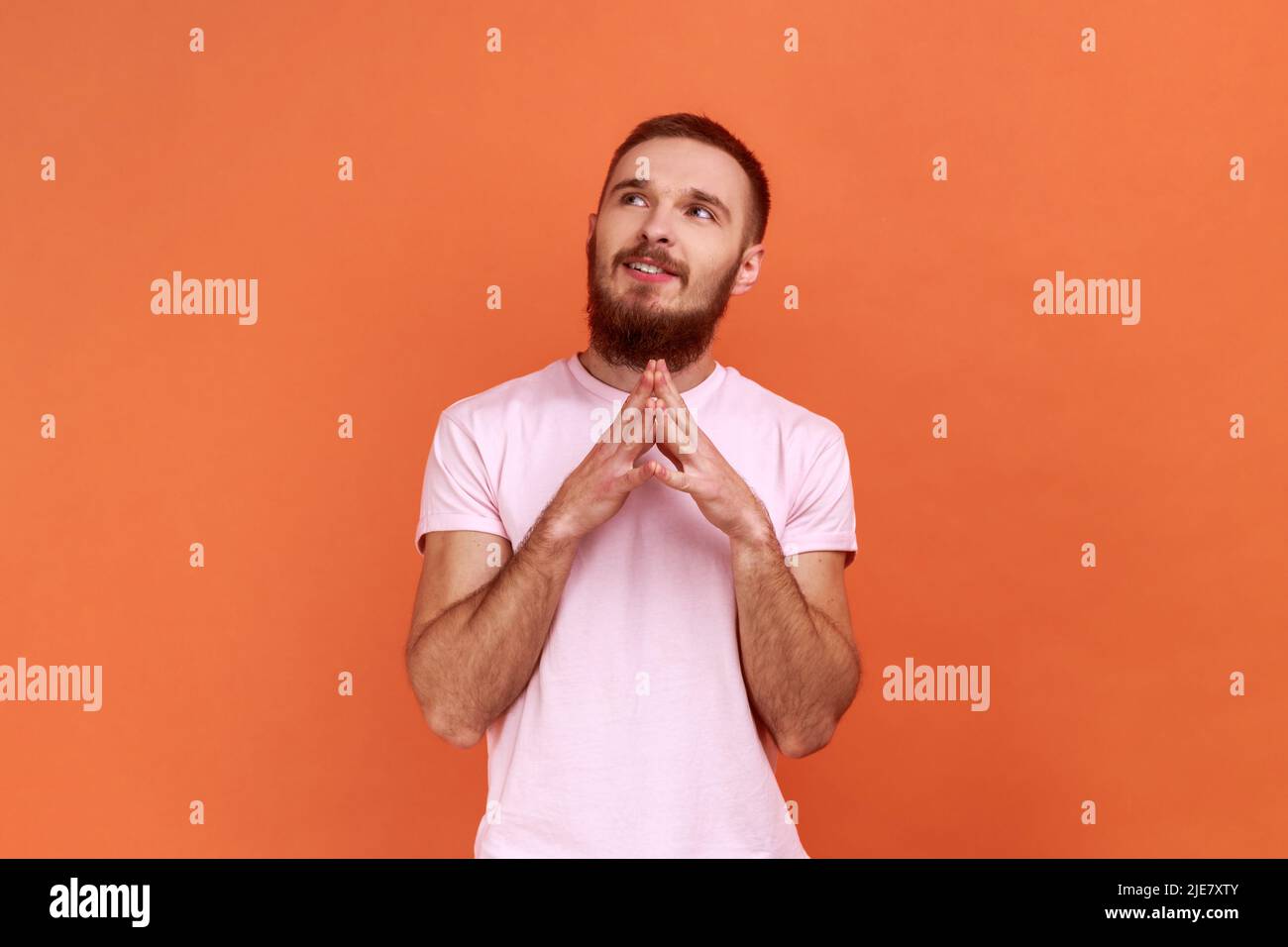 Portrait of bearded man thinking over plans for future, thoughtful guy dreaming about good job, looking away, wearing pink T-shirt. Indoor studio shot isolated on orange background. Stock Photo
