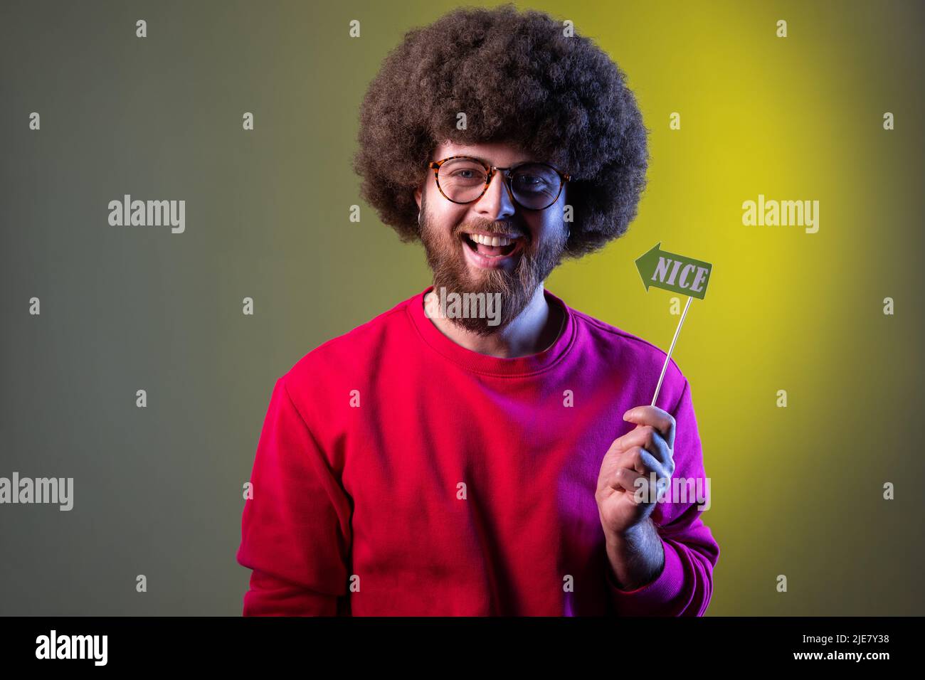 Happy positive hipster man with Afro hairstyle having event, holding party props with nice word in hands, wearing red sweatshirt. Indoor studio shot isolated on colorful neon light background. Stock Photo