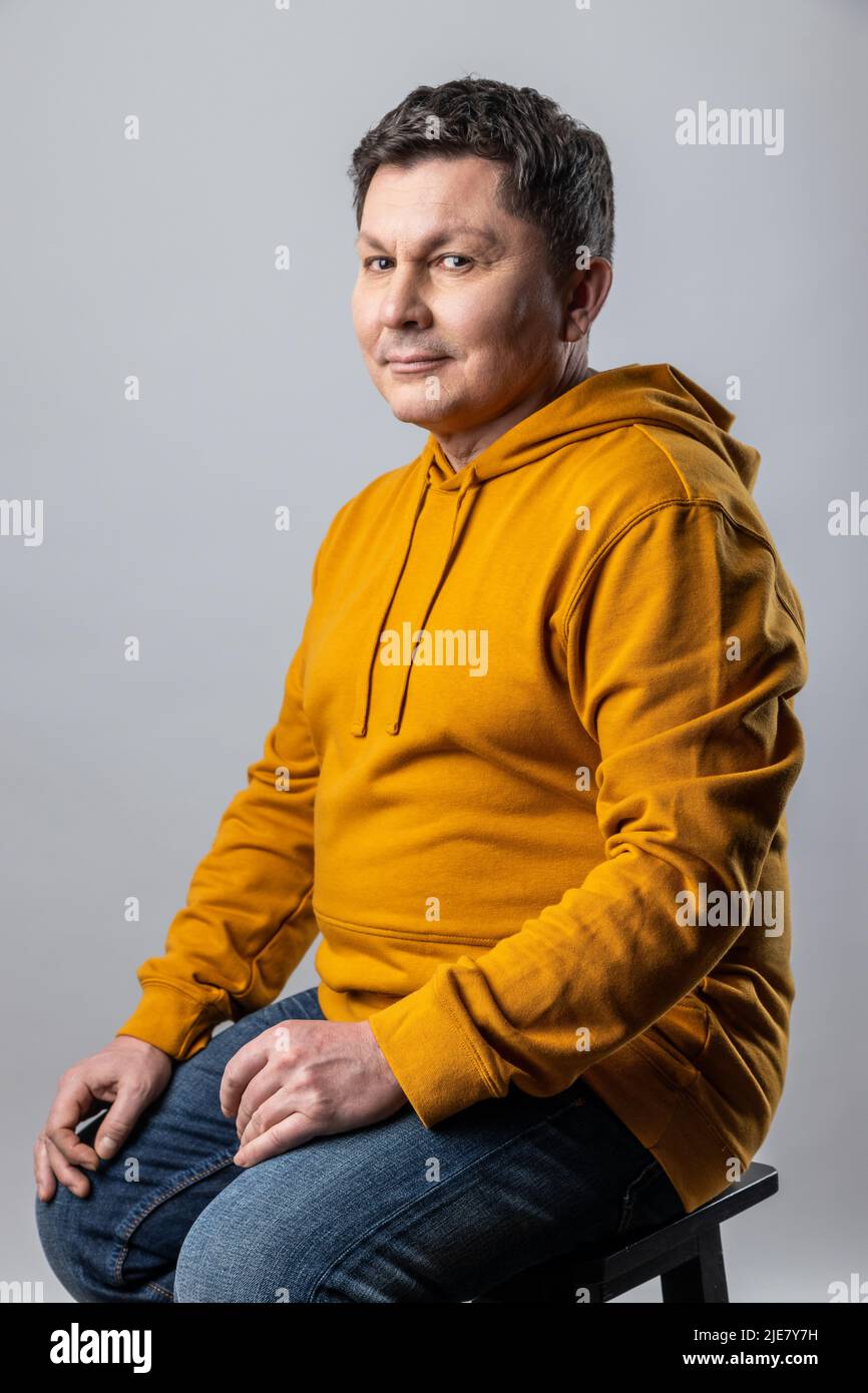 Portrait of attractive middle aged man looking at camera, sitting on chair and expressing positive emotions, wearing urban style hoodie. Indoor studio shot isolated on gray background. Stock Photo