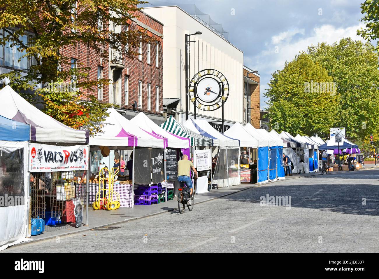 Shopping high street view pavement roadside market stalls in Brentwood Essex cyclists pedalling along cobble stone road clock wrong time England UK Stock Photo