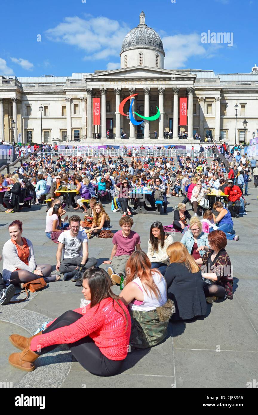 Tourists & visitors sit on paving no space at popular coloured picnic tables sunny day Trafalgar Square 2012 London Paralympic Games logo England UK Stock Photo