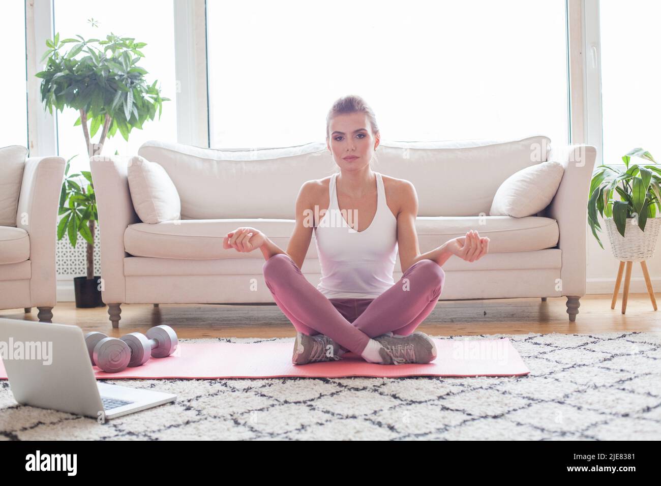 Fit sporty healthy woman sitting on mat, doing breathing exercises, watching online yoga class on laptop computer. Healthy people and self motivation Stock Photo