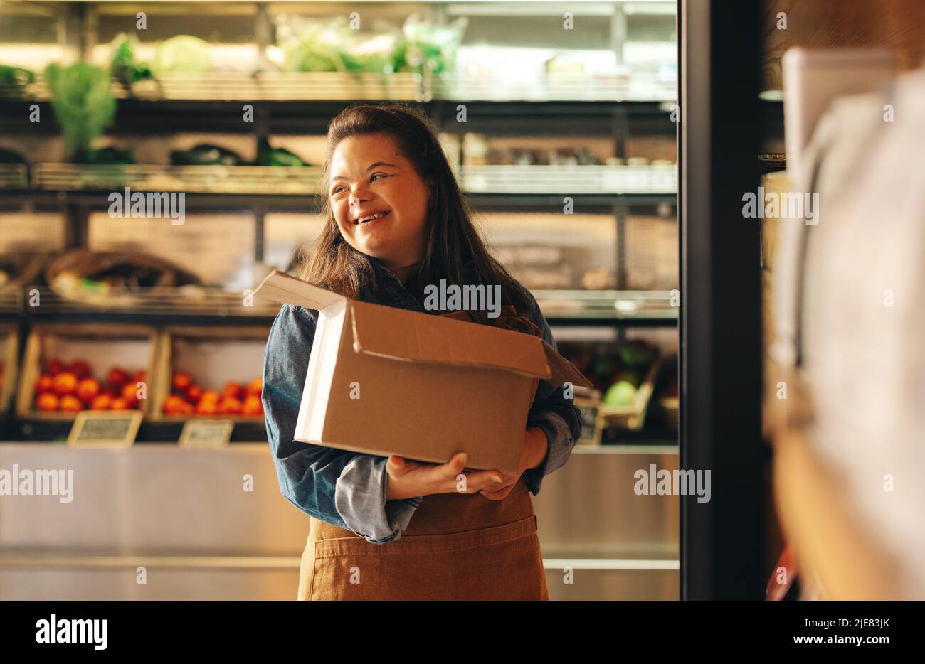Woman with Down syndrome smiling happily while working as a shopkeeper in a grocery store. Empowered woman with an intellectual disability restocking Stock Photo