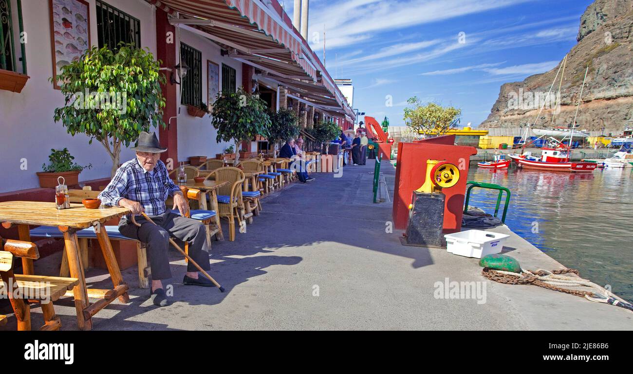 Old local man sitting on a bench at a fish restaurant, harbour of Puerto de Mogan, Grand Canary, Canary islands, Spain, Europe Stock Photo