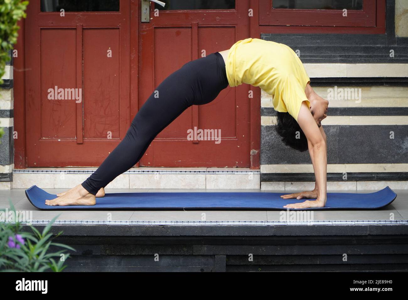 On his terrace, a young Asian girl with a stunning appearance is practicing Yoga while sporting a short haircut, a yellow shirt, and black leggings. S Stock Photo