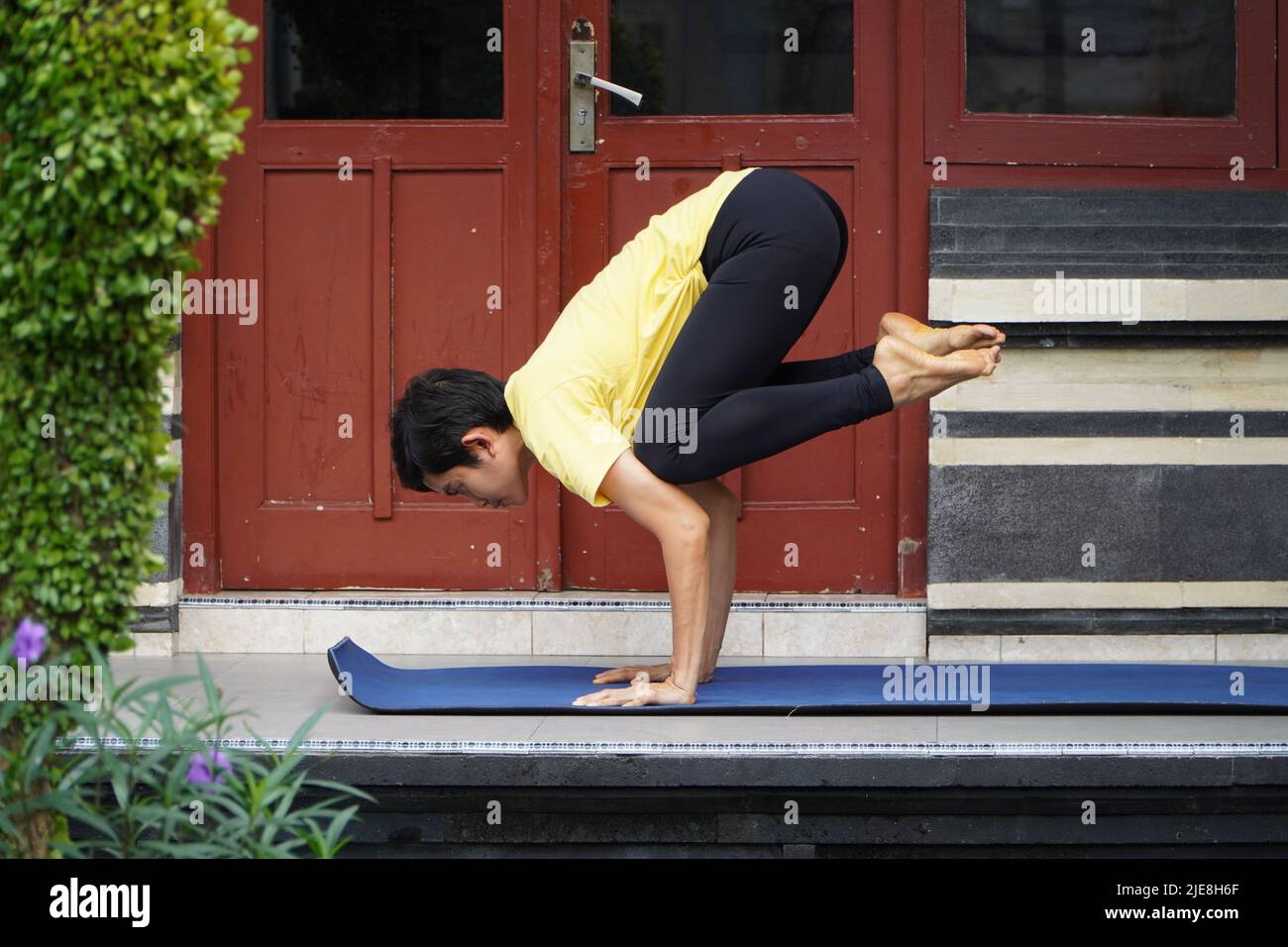 On his terrace, a young Asian girl with a stunning appearance is practicing Yoga while sporting a short haircut, a yellow shirt, and black leggings. S Stock Photo