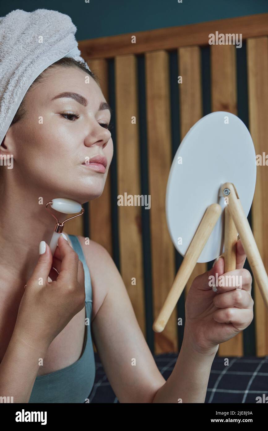Young woman using jade facial roller for face and neck massage sitting