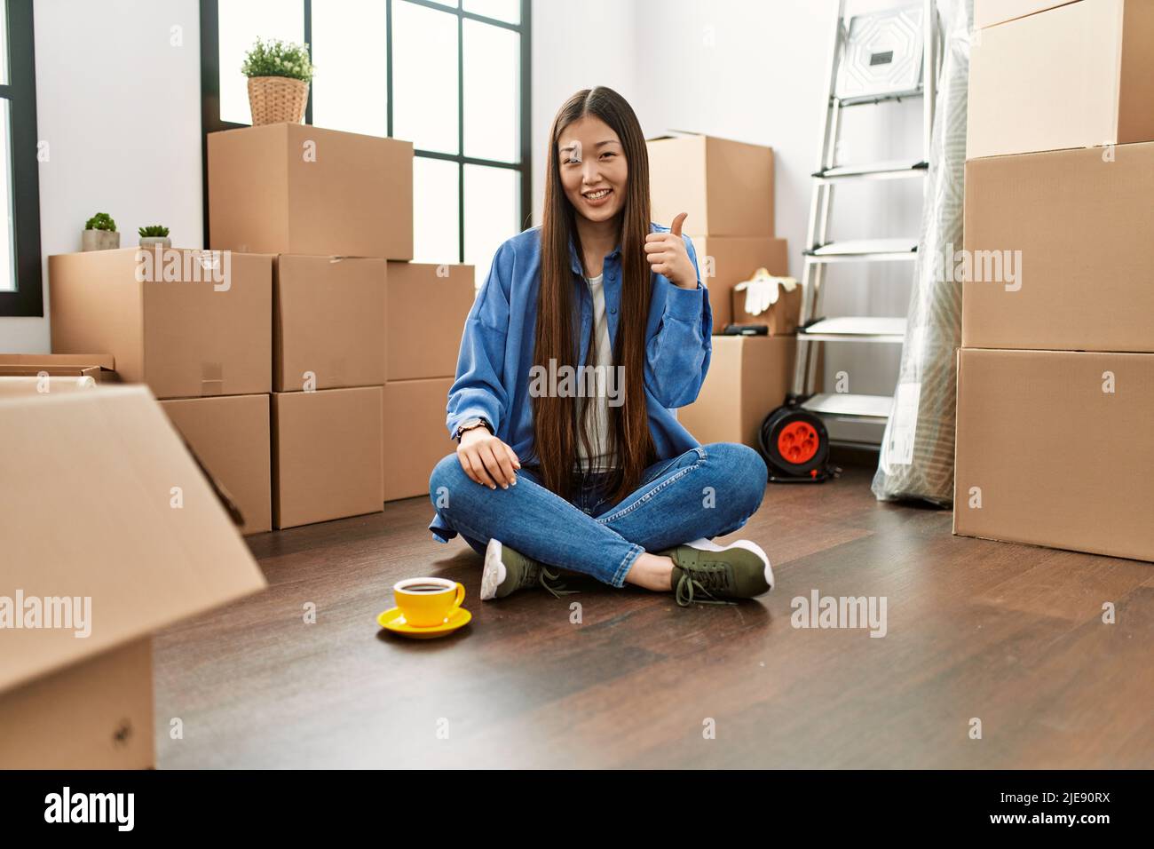 Young chinese girl sitting on the floor at new home doing happy thumbs ...