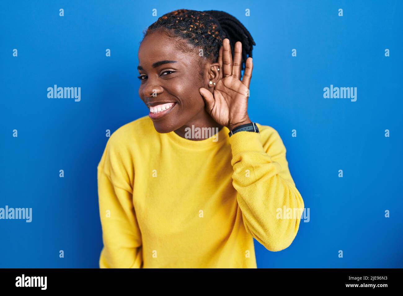 Beautiful black woman standing over blue background smiling with hand over ear  listening an hearing to rumor or gossip. deafness concept Stock Photo -  Alamy