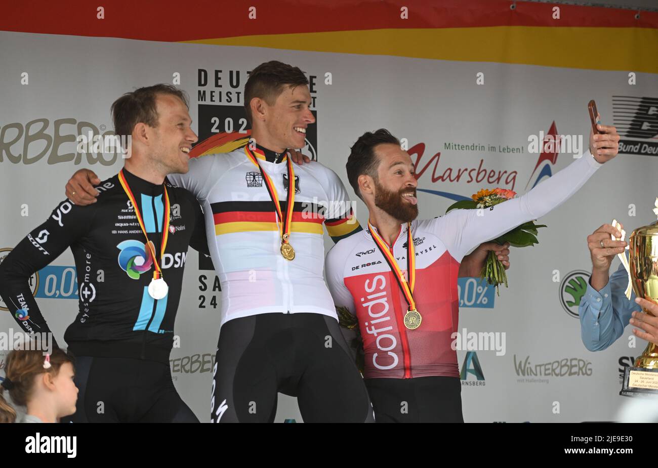 Winterberg, Germany. 26th June, 2022. Cycling: German Championship: Arnsberg - Winterberg (210.00 km): Third-placed Simon Geschke (r) from Team Cofidis takes a selfie with winner Nils Politt (M) from Bora - hansgrohe and second-placed Nikias Arndt from Team DSM after the award ceremony, sticking his tongue out. In front, Arndt's godchild, whom he had taken with him to the podium. Credit: Bernd Thissen/dpa/Alamy Live News Stock Photo