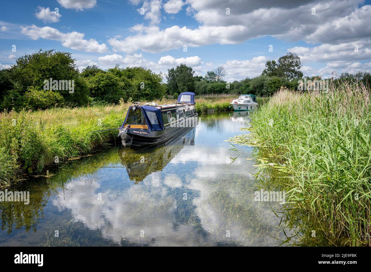 Wicken Cambridgeshire, 26th June 2022. Boats moored at Wicken Lode near Wicken Fen nature reserve with clouds reflected in the still waters. A beautiful summer day in the Cambridgeshire Fens, sun, blue skies and fluffy clouds skudding by in the breeze in the flat landscape. Credit: Julian Eales/Alamy Live News Stock Photo