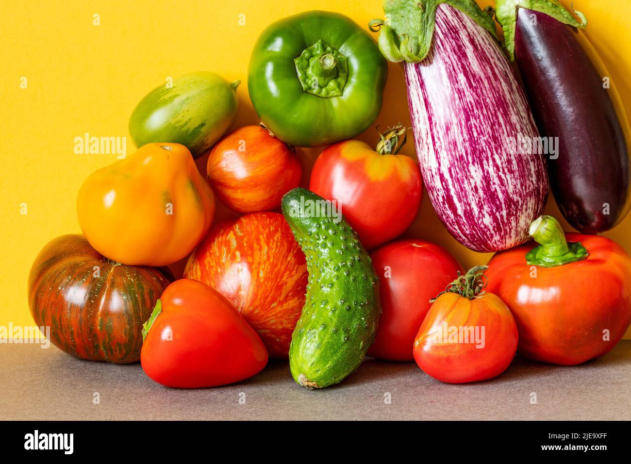 Organic vegetables still life scene. Farm aubergine eggplants, tomatoes of various grade, bell peppers and cucumber on a yellow gray background. Stock Photo