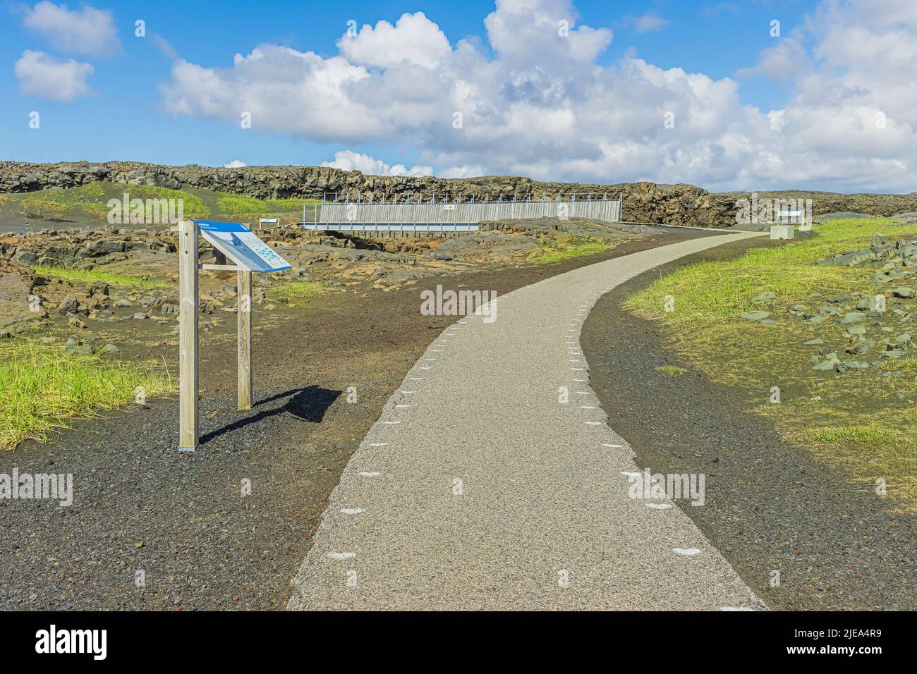 Pathway with a sign pointing to a metal bridge on the Reykjanes Peninsula of Iceland. Lava rocks and sand with green grass between the American and Eu Stock Photo