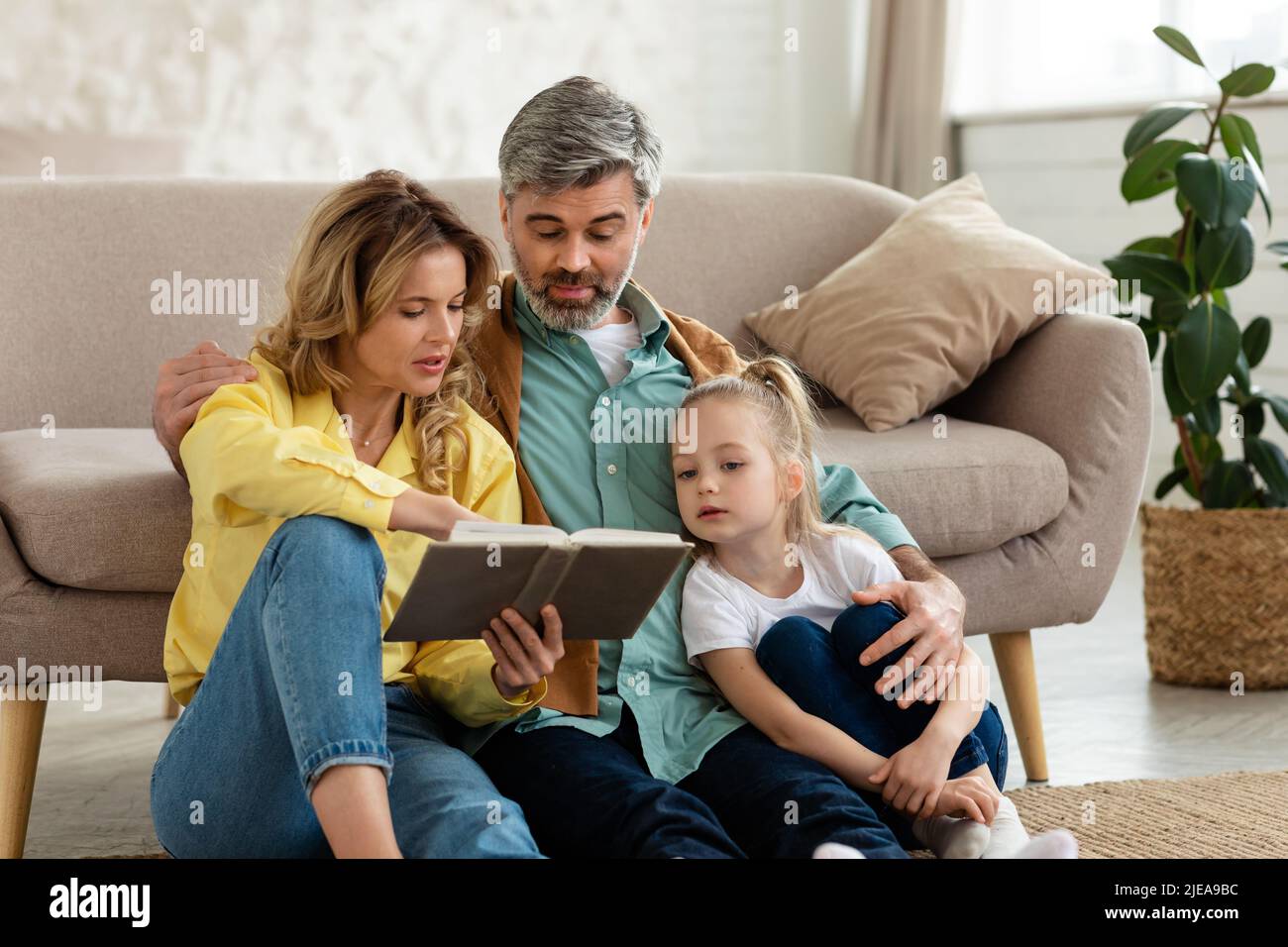 Family Reading Book Hugging Bonding Sitting Near Couch At Home Stock Photo