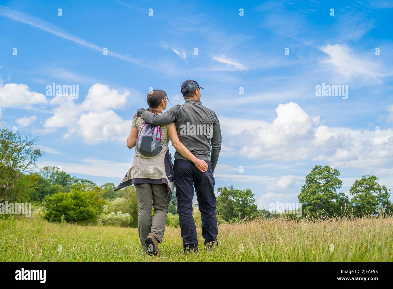 Kidderminster, UK. 26th June, 2022. UK weather: sunny weather and warm temperatures entice walkers for a stroll in the Worcestershire countryside. A couple, arms around each other, walk through a country meadow, stopping to enjoy the nature around them, with blue skies above. Credit: Lee Hudson/Alamy Live News Stock Photo