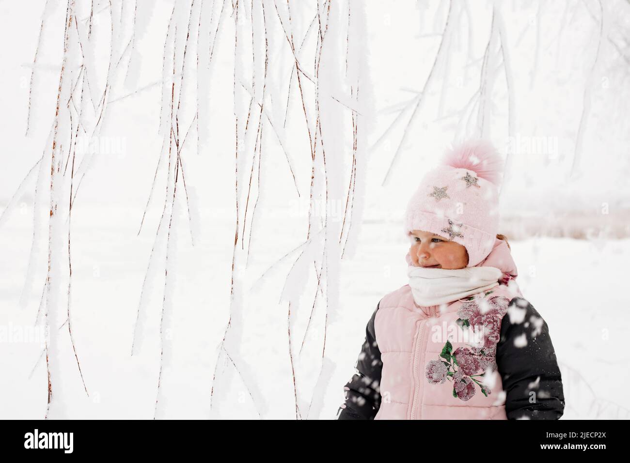 Tree branches covered with snow with blissful girl with rosy cheeks looking away dressed in warm winter clothes with snowy field in background. Winter Stock Photo