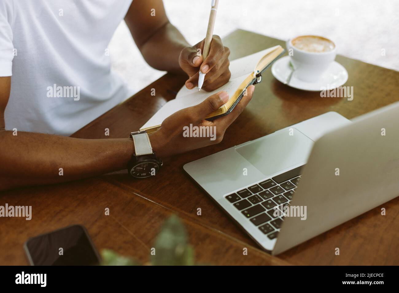 Unrecognizable cropped multicultural man using laptop, taking notes in workbook, drink coffee during internship study Stock Photo