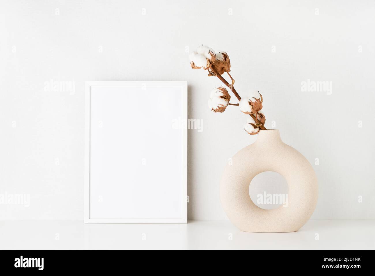 White frame mockup and round ceramic vase with a cotton branch on a white table by the light wall. Copy space for text Stock Photo