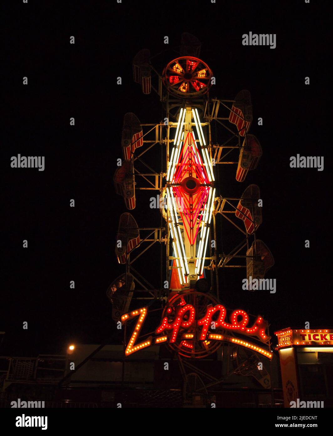 An old fashioned carnival ride and vintage ticket booth illuminated in neon and marquee style lighting at night against a black sky. Stock Photo
