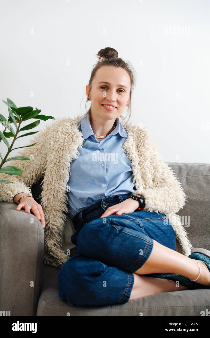 Comfortable confident 30 year old woman sitting on a couch, looking at camera. She has her hair in a knot, wearing blue shirt, jeans and a fashionable Stock Photo