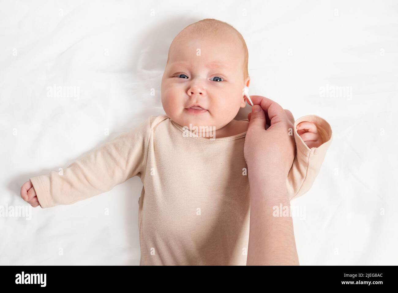 Hand of mother cleans ears of baby girl lying on white sheet with