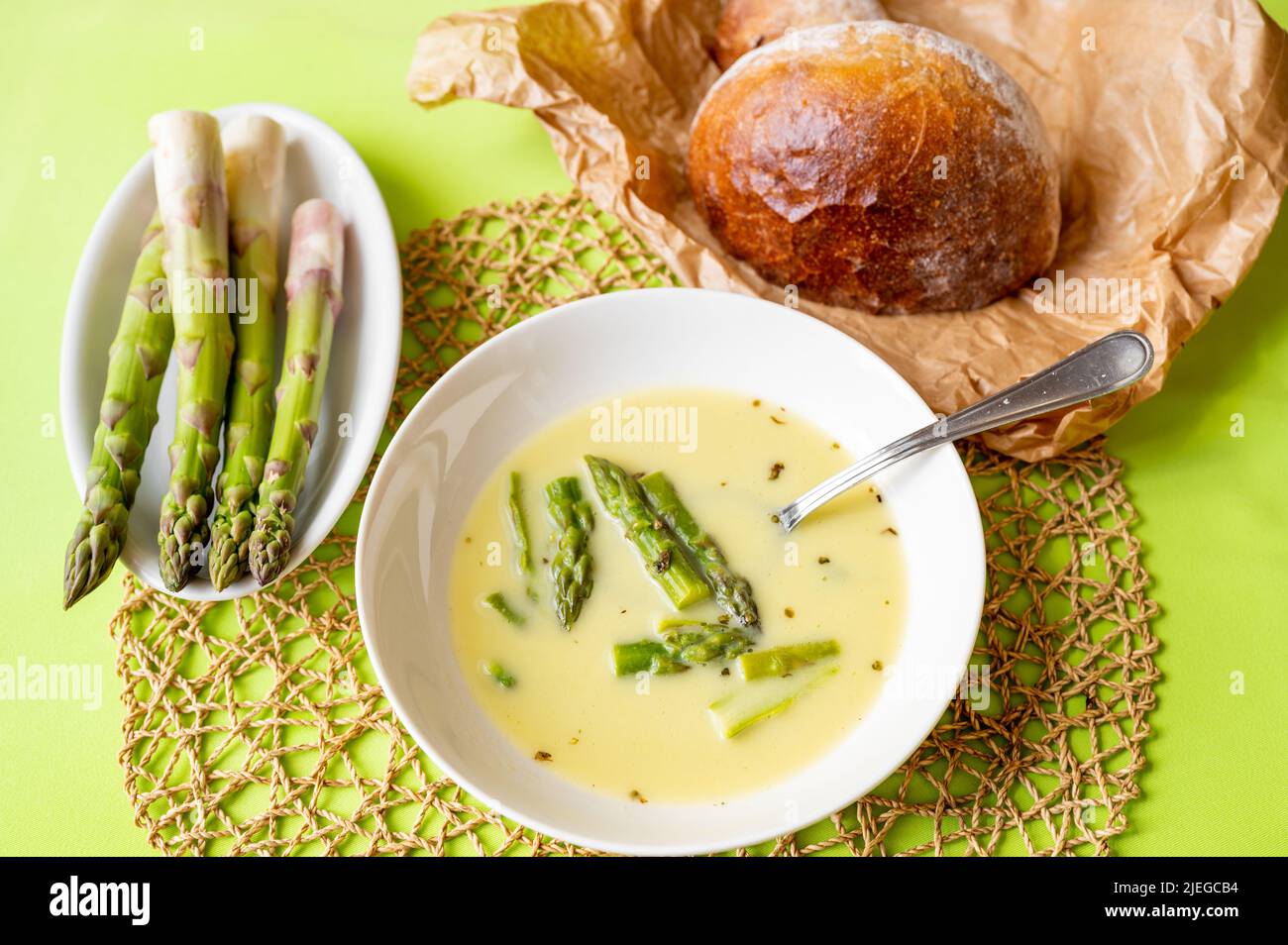 Asparagus soup in plate, boiled asparagus on bowl and loaf of bread on paper on green background. Stock Photo