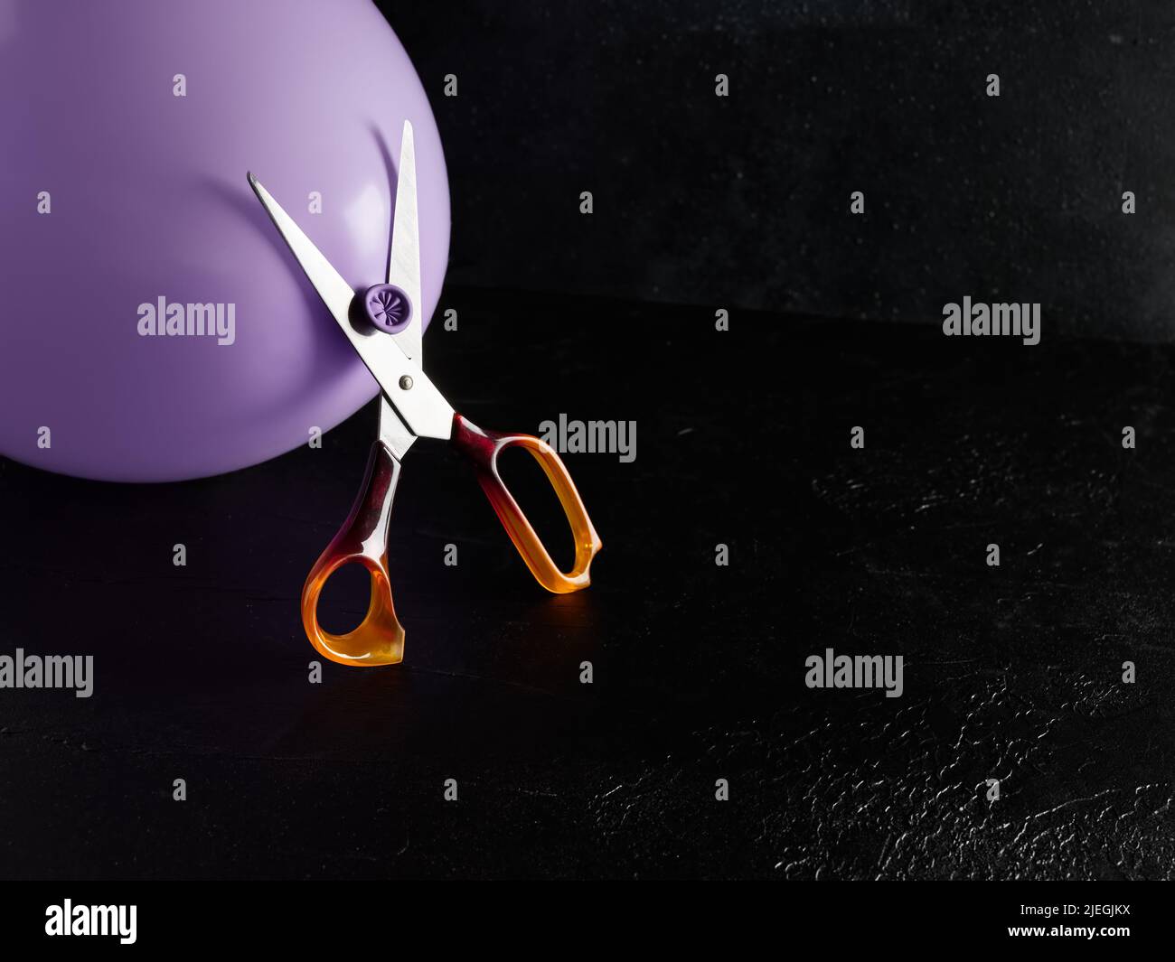 Scissors cutting the purple balloon against moody dark black background. Risk, insurance, danger or emotional pressure concepts Stock Photo