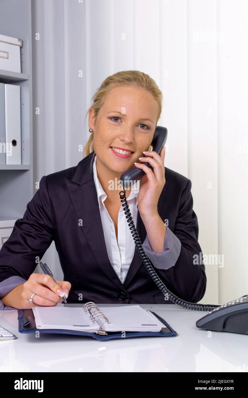 Eine freundliche Frau telefoniert an ihrem Schreibtisch im B¸ro und notiert Termine im Kalender Stock Photo